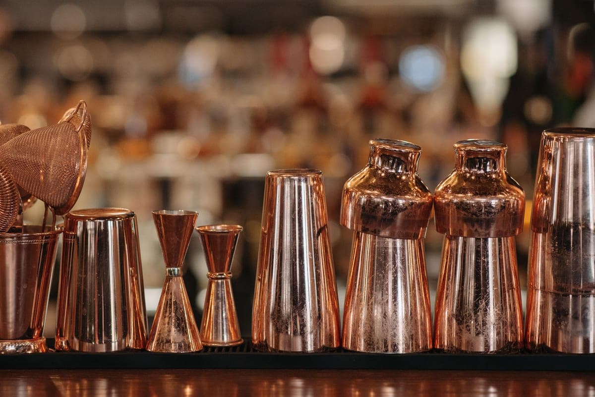 A cocktail shaker, jigger and strainer laid out on a work surface