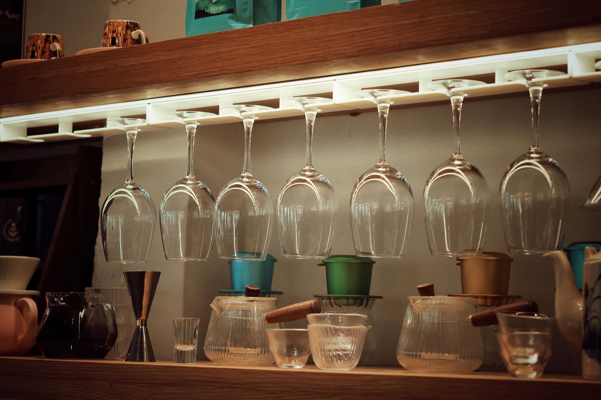 Stemmed glasses hanging inverted from a rack beneath a wood shelf, with bar tools below