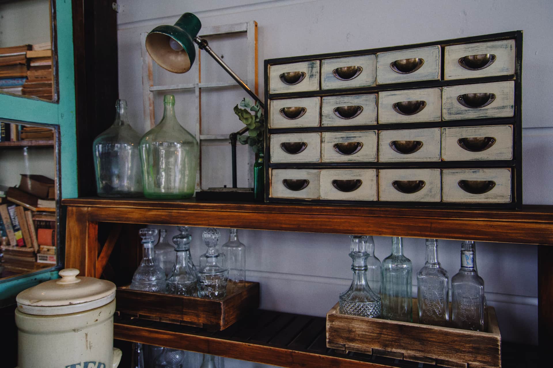 A vintage cabinet in the corner of a room with glass bottles on a wooden shelf above it