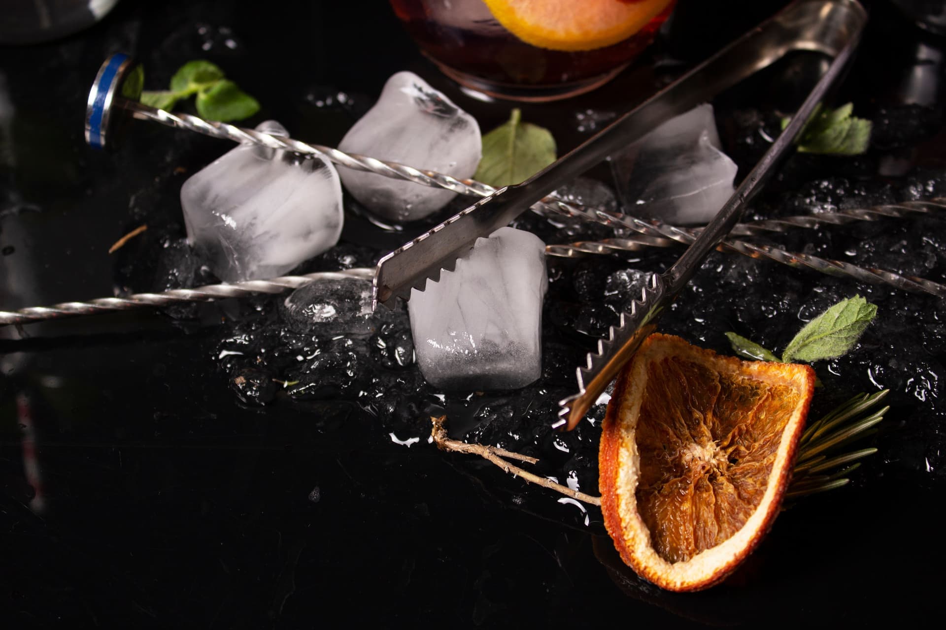 Ice cubes, a sliced orange and cocktail tools arranged on a dark surface